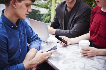 Drei Personen arbeiten an einem Tisch im Freien, zwei halten Handys in der Hand, einer tippt auf einem Laptop. - MINF05869