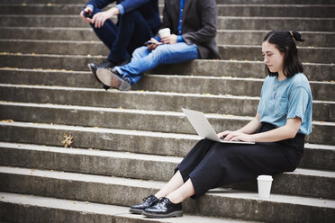 Eine junge Frau, die auf einer Treppe im Freien sitzt und an einem Laptop arbeitet, mit zwei Männern im Hintergrund. - MINF05874