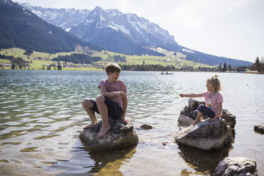 Österreich, Tirol, Walchsee, Bruder und Schwester sitzen auf Felsbrocken im See und spielen mit Stöcken - JLOF00159