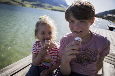 Österreich, Tirol, Walchsee, Porträt von Bruder und Schwester, die auf einem Steg am See sitzen und Eistüten essen - JLOF00180