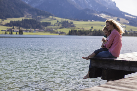 Österreich, Tirol, Walchsee, glückliche Mutter und Tochter sitzen auf einem Steg am See, lizenzfreies Stockfoto