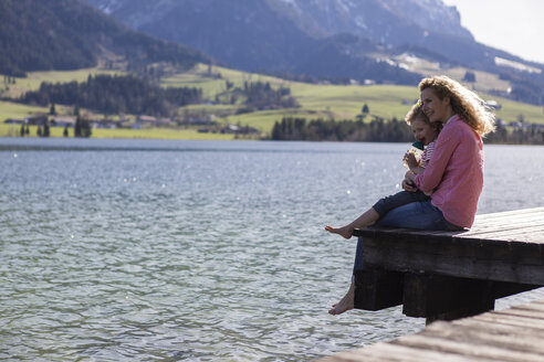 Austria, Tyrol, Walchsee, happy mother and daughter sitting on a jetty at the lake - JLOF00183