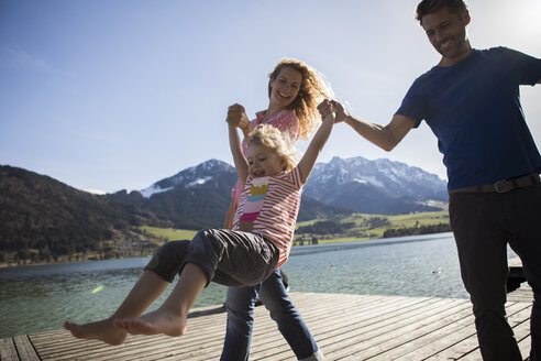 Österreich, Tirol, Walchsee, glückliche Eltern schaukeln ihre Tochter am See - JLOF00186