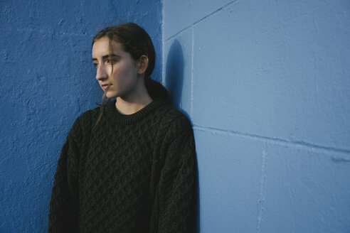Porträt eines Mädchens mit braunen Haaren im Pferdeschwanz und schwarzem Strickpullover, das an einer blauen Wand lehnt. - MINF05886