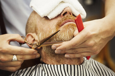 Ein Kunde sitzt auf dem Friseurstuhl, hat ein heißes Handtuch im Gesicht und ein Friseur stutzt ihm den Bart. - MINF05901