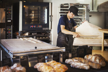 Ein Mann arbeitet in einer Bäckerei und bereitet ein großes Tablett mit Teig für Brötchen vor, im Hintergrund ein Ofen. - MINF05919