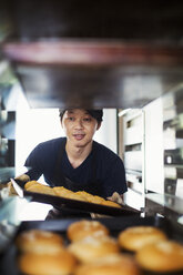 Ein Mann arbeitet in einer Bäckerei und legt große Tabletts mit frisch gebackenen Brötchen auf einen Wagen. - MINF05920