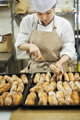 Frau mit Kochmütze und Schürze arbeitet in einer Bäckerei und schneidet frisch gebackene Brötchen auf großen Tabletts auf. - MINF05921