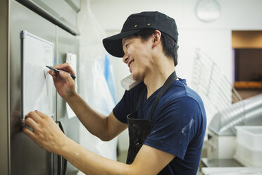 Mann arbeitet in einer Bäckerei, trägt Baseballkappe und Schürze, schreibt Notizen auf eine kleine Tafel, telefoniert und lächelt. - MINF05922