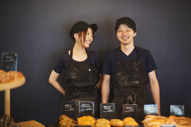 Smiling man and woman wearing baseball cap and apron standing in a bakery, trays with freshly baked goods. - MINF05923