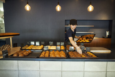 Man working in a bakery, placing freshly baked croissants and cakes on large trays on a counter. - MINF05924