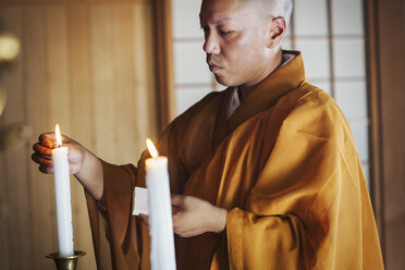 Seitenansicht eines buddhistischen Mönchs mit rasiertem Kopf und goldenem Gewand, der in einem Tempel kniet und eine weiße Kerze anzündet. - MINF05955