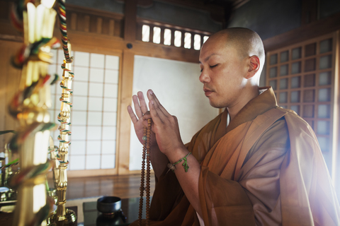Seitenansicht eines buddhistischen Mönchs mit rasiertem Kopf und goldenem Gewand, der in einem Tempel kniet und eine Mala hält, mit geschlossenen Augen., lizenzfreies Stockfoto
