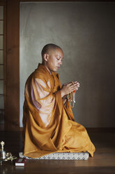 Side view of Buddhist monk with shaved head wearing golden robe kneeling indoors in a temple, holding mala, praying. - MINF05957