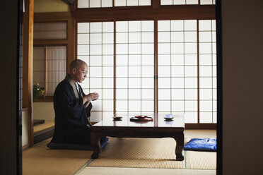 Seitenansicht eines buddhistischen Mönchs mit rasiertem Kopf und schwarzer Robe, der in einem Haus an einem Tisch kniet und eine Schale mit Tee hält. - MINF05958