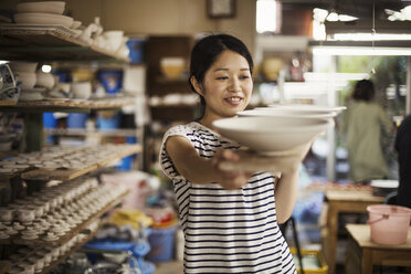 Woman standing in a workshop, carrying long wooden tray with porcelain bowls. - MINF05960