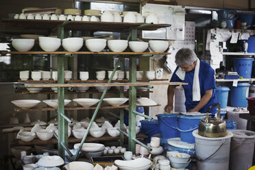 Man standing in a Japanese porcelain workshop with shelves of various porcelain bowls. - MINF05963