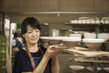Frau steht in einer japanischen Porzellanwerkstatt und trägt ein langes Holztablett mit Schalen. - MINF05964