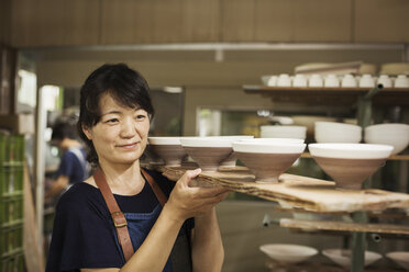 Woman standing in a Japanese porcelain workshop, carrying long wooden tray with bowls. - MINF05964