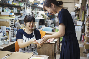 Two smiling women sitting and standing in a workshop, looking at Japanese porcelain bowl. - MINF05965