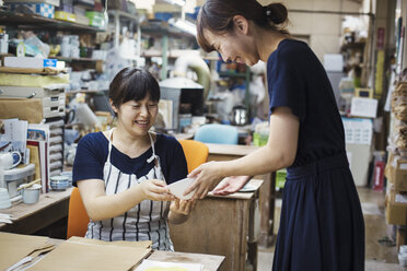 Zwei lächelnde Frauen sitzen und stehen in einer Werkstatt und betrachten eine japanische Porzellanschale. - MINF05965