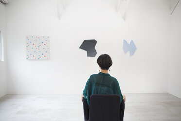 Rückansicht einer Frau mit kurzen schwarzen Haaren und grünem Hemd, die in einer Kunstgalerie sitzt und sich moderne Gemälde ansieht. - MINF05973