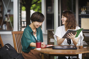 Zwei Frauen mit schwarzen Haaren und grün-weißem Hemd sitzen an einem Tisch in einem Straßencafé, halten ein digitales Tablet in der Hand und lächeln. - MINF05981