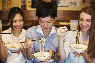 Drei lächelnde Menschen sitzen nebeneinander an einem Tisch in einem Restaurant und essen mit Stäbchen aus Schüsseln. - MINF05992