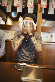 Mann mit tätowierten Armen sitzt an einem Tisch in einem Restaurant und wischt sich mit einem nassen Handtuch das Gesicht ab. - MINF05998