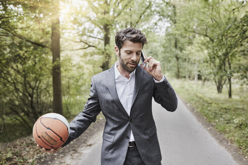 Geschäftsmann mit Basketball, der auf einer Landstraße mit seinem Smartphone spricht - RORF01363
