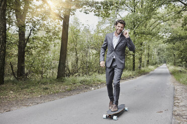 Businessman riding skateboard and talking on smartphone on rural road - RORF01368