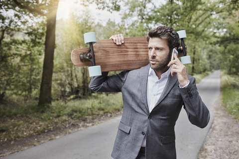Geschäftsmann mit Skateboard, der auf einer Landstraße mit seinem Smartphone spricht, lizenzfreies Stockfoto