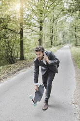 Businessman walking with skateboard and smartphone on rural road - RORF01373