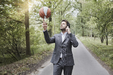 Businessman balancing basketball and talking on smartphone on rural road - RORF01375
