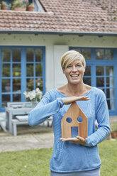 Portrait of smiling woman standing in front of her home holding house model - RORF01406