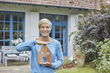 Portrait of smiling woman standing in front of her home holding house model - RORF01408