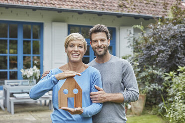 Portrait of smiling couple standing in front of their home holding house model - RORF01409
