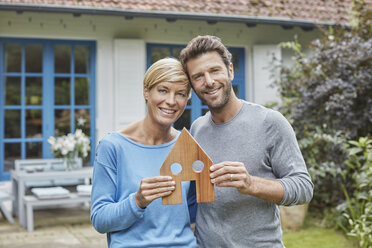 Portrait of smiling couple standing in front of their home holding house model - RORF01410