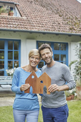 Portrait of smiling couple standing in front of their home holding house model - RORF01412