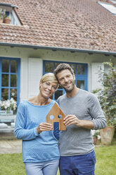 Portrait of smiling couple standing in front of their home holding house model - RORF01413