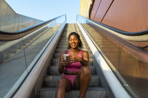 junge Frau sitzt auf der Rolltreppe und benutzt ihr Smartphone, lizenzfreies Stockfoto