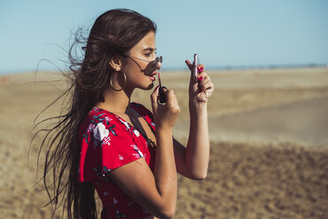 Teenage girl applying lip gloss on the beach - ACPF00160