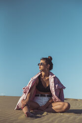 Teenage girl sitting on beach dune against blue sky - ACPF00169