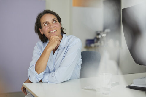 Porträt einer lächelnden reifen Geschäftsfrau, die am Schreibtisch in einem Büro sitzt und nach oben schaut - PNEF00847