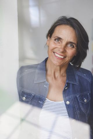 Porträt einer lächelnden reifen Frau hinter einer Fensterscheibe, lizenzfreies Stockfoto