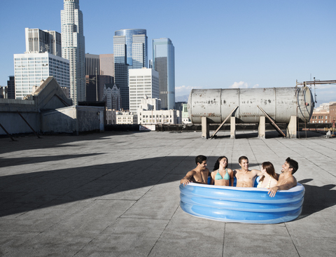 Eine Gruppe von Freunden, Männer und Frauen, sitzt in einem kleinen aufblasbaren Wasserbecken auf einem städtischen Dach und kühlt sich ab., lizenzfreies Stockfoto