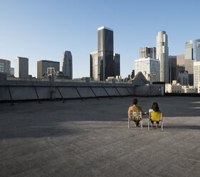 Ein Paar, Mann und Frau, sitzen in Liegestühlen auf einem Dach mit Blick auf die Wolkenkratzer der Stadt. - MINF06050