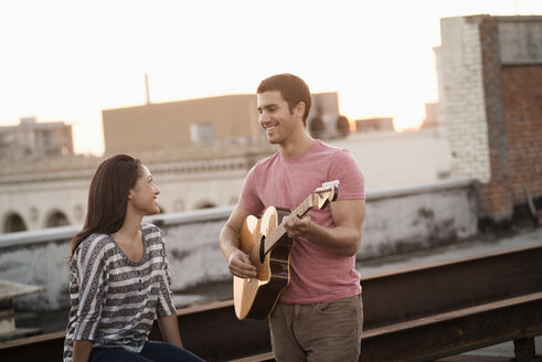 Ein Mann spielt einer Frau auf einer Dachterrasse mit Blick auf eine Stadt in der Abenddämmerung Gitarre vor. - MINF06056