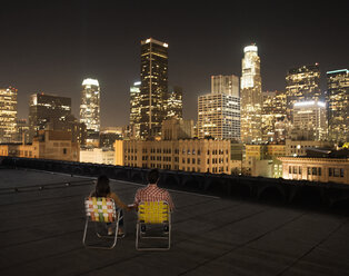 Ein Paar auf einer Dachterrasse mit Blick auf das nächtliche Los Angeles, das Seite an Seite sitzt und über die Stadt blickt. - MINF06068