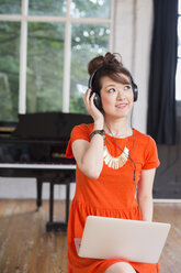 Young woman wearing headphones, standing in a rehearsal studio, listening to music, balancing a laptop computer on her knee. - MINF06122
